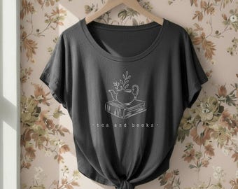 May include: A dark gray short-sleeve t-shirt with a wide neckline and a knotted hem. The front features a white line-art graphic of a teapot resting on a book with a sprig of leaves, accompanied by the text "tea and books" in lowercase letters.