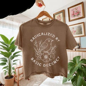 May include: A brown t-shirt with the text "RADICALIZED BY BASIC DECENCY" in a white, stylized font. A white illustration of a bird perched on flowers and leaves is centered below the text. The shirt is hanging on a wooden hanger.
