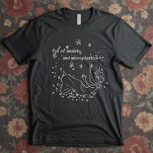 May include: Dark gray t-shirt with a white graphic of a person floating in a circle of dots and flowers. The text "full of anxiety and microplastics" is above the figure.