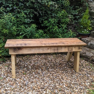 May include: A rustic wooden bench made from reclaimed wood. The bench has a natural wood finish and is sitting on a gravel surface.