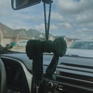 May include: A dark green, handmade crochet car charm suspended from a car's rearview mirror. The charm is shaped like a bow tie with two long, dangling sections. The background shows a suburban street and a cloudy sky.