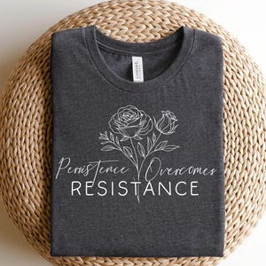 May include: Dark gray t-shirt with a white floral design and the words "Persistence Overcomes Resistance". The shirt is folded and placed on a woven, circular surface.