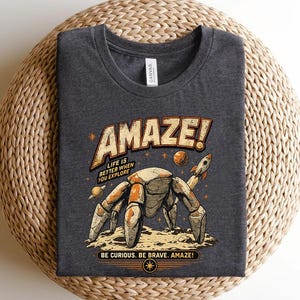 May include: A dark heather gray t-shirt featuring a graphic of a robotic crab on a lunar surface with a rocket and planets. The text reads AMAZE! LIFE IS BETTER WHEN YOU EXPLORE and BE CURIOUS. BE BRAVE. AMAZE! in a retro, distressed orange and cream font.