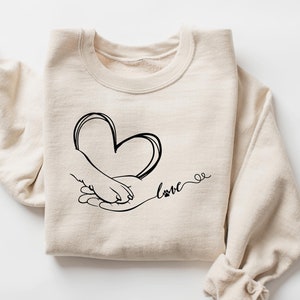 Könnte beinhalten: Ein beiger Pullover mit einer schwarzen Grafik einer menschlichen Hand und einer Pfote, die ein Herz mit dem Wort "love" und einer Pfote darunter halten.