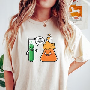 Puede incluir: Camiseta beige con un diseño de dibujos animados de dos matraces de laboratorio. Un matraz verde señala a un matraz naranja con una expresión preocupada. El texto en un bocadillo dice: "Creo que estás exagerando."