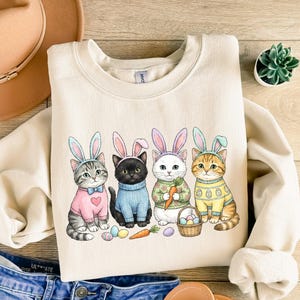 May include: Cream-colored sweatshirt featuring a graphic of four cats dressed for Easter, each wearing bunny ears and sweaters. The cats are surrounded by Easter eggs, a carrot, and a basket. The sweatshirt is paired with blue jeans and a tan hat.