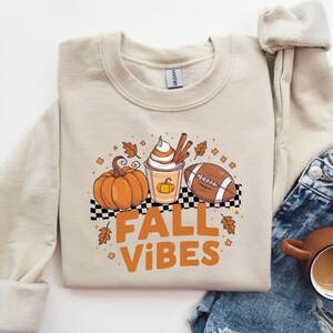May include: A beige sweatshirt with a graphic design of a pumpkin, a cup of coffee with whipped cream, and a football. The text "Fall Vibes" is printed below the graphic.
