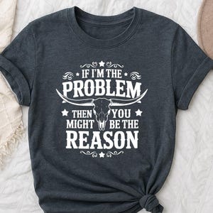 Może przedstawiać: Ciemnoszara koszulka z białym napisem "IF I'M THE PROBLEM THEN YOU MIGHT BE THE REASON". Projekt zawiera czaszkę byka i gwiazdy. Koszulka ma krótkie rękawy i jest wykonana z miękkiego materiału.