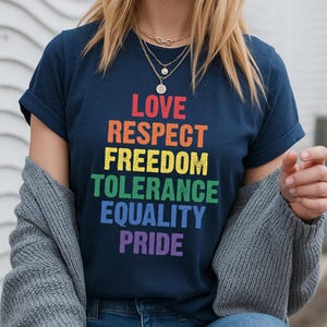 Puede incluir: Camiseta azul marino con las palabras "LOVE, RESPECT, FREEDOM, TOLERANCE, EQUALITY, PRIDE" en colores arcoíris. La camiseta se combina con vaqueros azules y una rebeca gris. El tema es el orgullo LGBTQ+.