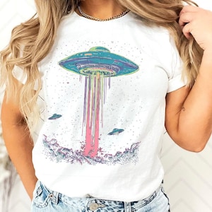 May include: White t-shirt with a vibrant graphic of a UFO. The design shows a blue and green UFO with a beam of pink and purple light. There are smaller UFOs in the background. This casual t-shirt is suitable for everyday wear.