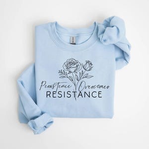 May include: A light blue sweatshirt with a floral design and the words "Persistence Overcomes Resistance" in black script. The design features two roses and leaves. The sweatshirt is folded neatly.