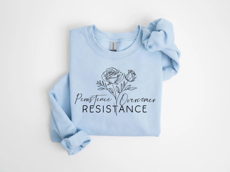 May include: A light blue sweatshirt with a floral design and the words "Persistence Overcomes Resistance" in black script. The design features two roses and leaves. The sweatshirt is folded neatly.