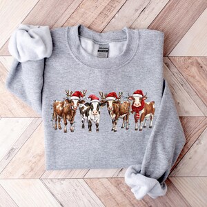 May include: A gray sweatshirt with a Christmas-themed design featuring four cows wearing Santa hats. The cows are brown and white with red and white Santa hats.