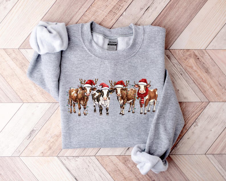 May include: A gray sweatshirt with a Christmas-themed design featuring four cows wearing Santa hats. The cows are brown and white with red and white Santa hats.
