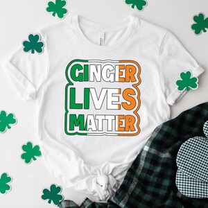 May include: White t-shirt with a green, white, and orange graphic that says "Ginger Lives Matter". The graphic is in the style of the Irish flag. The shirt is surrounded by green shamrocks.