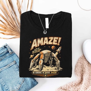 May include: A black t-shirt featuring a graphic of a robotic crab on a moon surface with planets and a rocket. The text reads AMAZE! LIFE IS BETTER WHEN YOU EXPLORE and BE CURIOUS. BE BRAVE. AMAZE! The design uses orange, tan, and white colors.