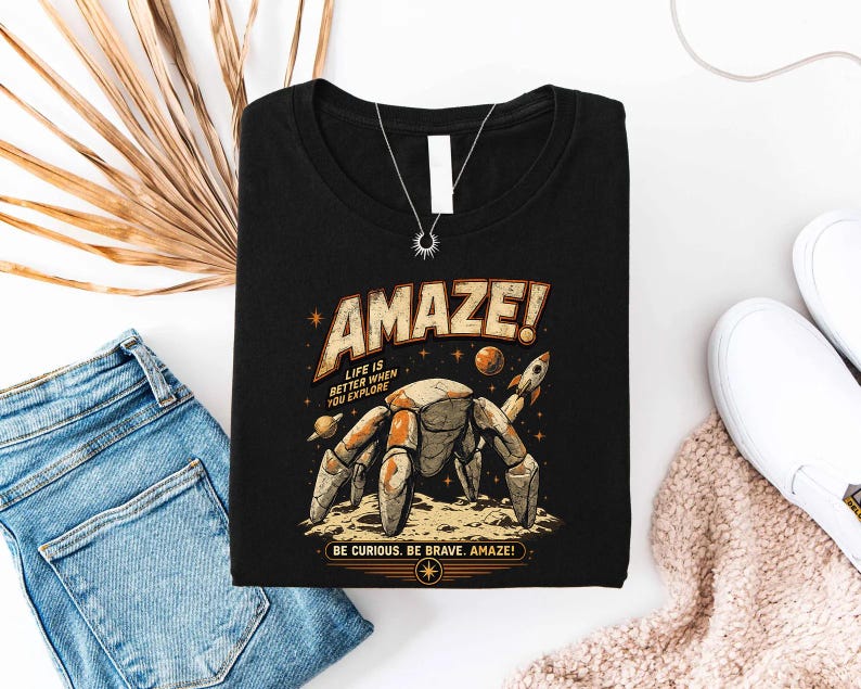 May include: A black t-shirt featuring a graphic of a robotic crab on a moon surface with planets and a rocket. The text reads AMAZE! LIFE IS BETTER WHEN YOU EXPLORE and BE CURIOUS. BE BRAVE. AMAZE! The design uses orange, tan, and white colors.