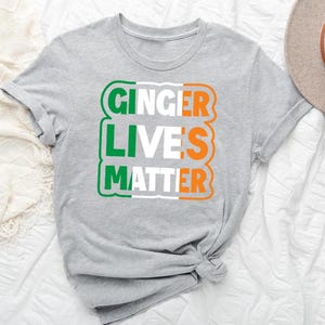 May include: Gray t-shirt with the text "Ginger Lives Matter" in white, green, and orange letters. The letters are arranged in a rectangle with a green, white, and orange color scheme, resembling the Irish flag.
