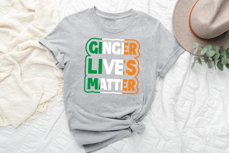 May include: Gray t-shirt with the text "Ginger Lives Matter" in white, green, and orange letters. The letters are arranged in a rectangle with a green, white, and orange color scheme, resembling the Irish flag.