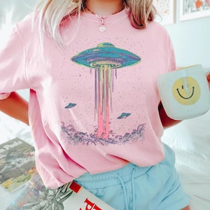 May include: Pink t-shirt with a colourful graphic of a UFO emitting a beam of light, and two smaller flying saucers. The shirt is worn with blue shorts. A white mug with a smiley face is held.