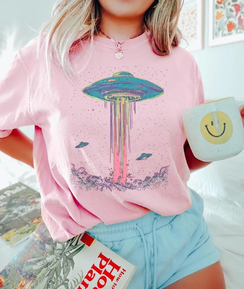 May include: Pink t-shirt with a colourful graphic of a UFO emitting a beam of light, and two smaller flying saucers. The shirt is worn with blue shorts. A white mug with a smiley face is held.