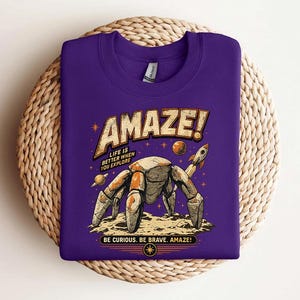May include: A purple crew neck t-shirt featuring a graphic of a robotic crab on a moon surface with a rocket, planets, and stars. The text reads "AMAZE! LIFE IS BETTER WHEN YOU EXPLORE" and "BE CURIOUS. BE BRAVE. AMAZE!" in a vintage-style font.