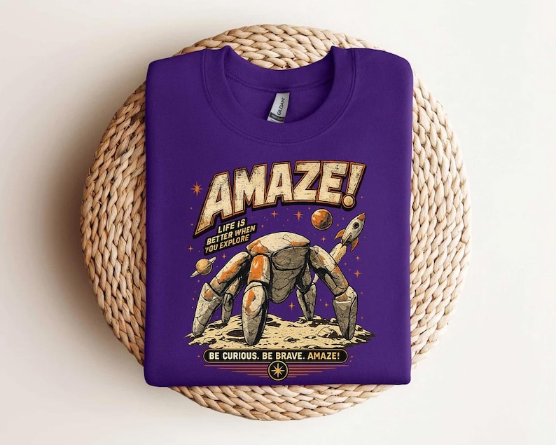 May include: A purple crew neck t-shirt featuring a graphic of a robotic crab on a moon surface with a rocket, planets, and stars. The text reads "AMAZE! LIFE IS BETTER WHEN YOU EXPLORE" and "BE CURIOUS. BE BRAVE. AMAZE!" in a vintage-style font.