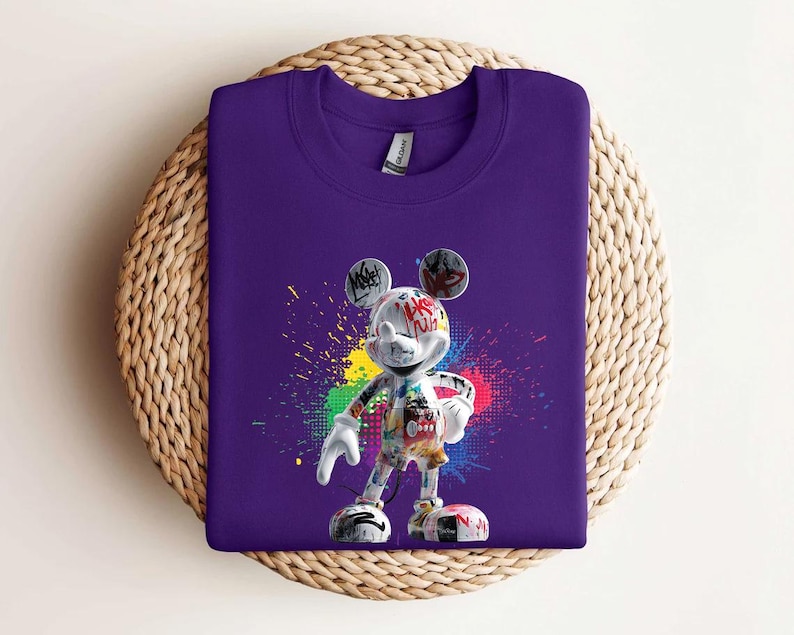 May include: A purple crew neck sweatshirt with a graphic of Mickey Mouse. The cartoon character is decorated with graffiti-style art and paint splatters. The sweatshirt is displayed on a woven, circular surface.