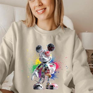 May include: A cream-coloured sweatshirt with a colourful graffiti-style Mickey Mouse graphic. The design includes vibrant paint splatters and text, creating a bold and artistic statement. The sweatshirt is a casual, comfortable garment.