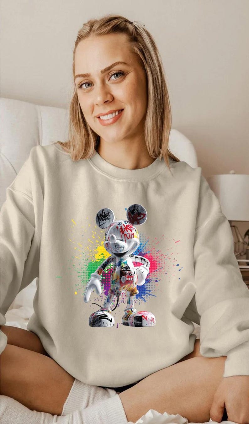May include: A cream-coloured sweatshirt with a colourful graffiti-style Mickey Mouse graphic. The design includes vibrant paint splatters and text, creating a bold and artistic statement. The sweatshirt is a casual, comfortable garment.