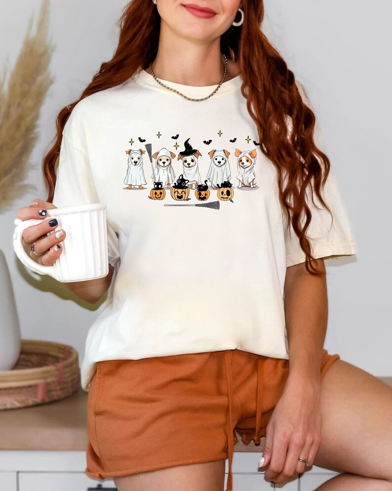 Op de afbeelding: Wit T-shirt met een afbeelding van vijf honden die verkleed zijn als geesten voor Halloween. De honden dragen heksenhoeden en houden bezems vast. Er zijn pompoenen en vleermuizen in het ontwerp.