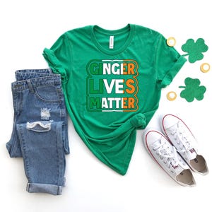 May include: Green t-shirt with the text "Ginger Lives Matter" in white, orange, and green letters. The shirt is styled with blue jeans and white sneakers.