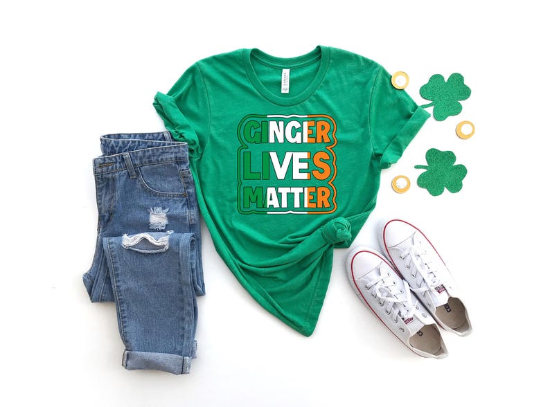 May include: Green t-shirt with the text "Ginger Lives Matter" in white, orange, and green letters. The shirt is styled with blue jeans and white sneakers.