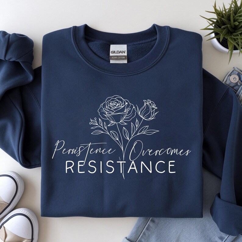 May include: Navy blue sweatshirt with a white floral design and the words "Persistence Overcomes Resistance." The sweatshirt is made of a soft material and is suitable for casual wear. The Gildan tag is visible.