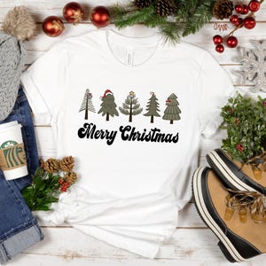 May include: White t-shirt with a graphic of five stylized Christmas trees and the text "Merry Christmas". The shirt is displayed on a white wooden surface with other Christmas decorations.