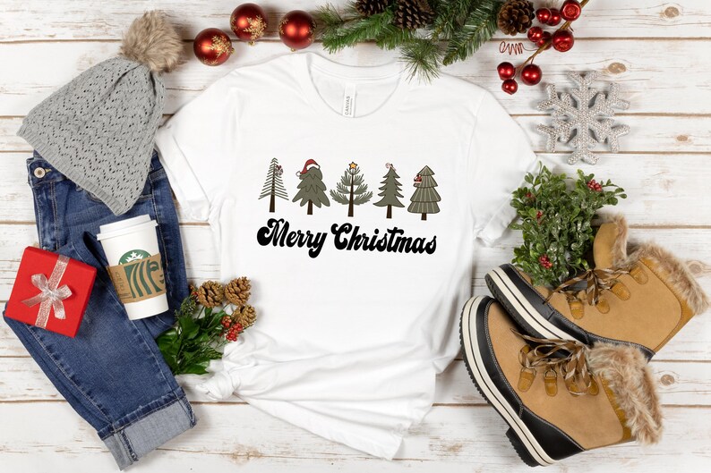 May include: White t-shirt with a graphic of five stylized Christmas trees and the text "Merry Christmas". The shirt is displayed on a white wooden surface with other Christmas decorations.