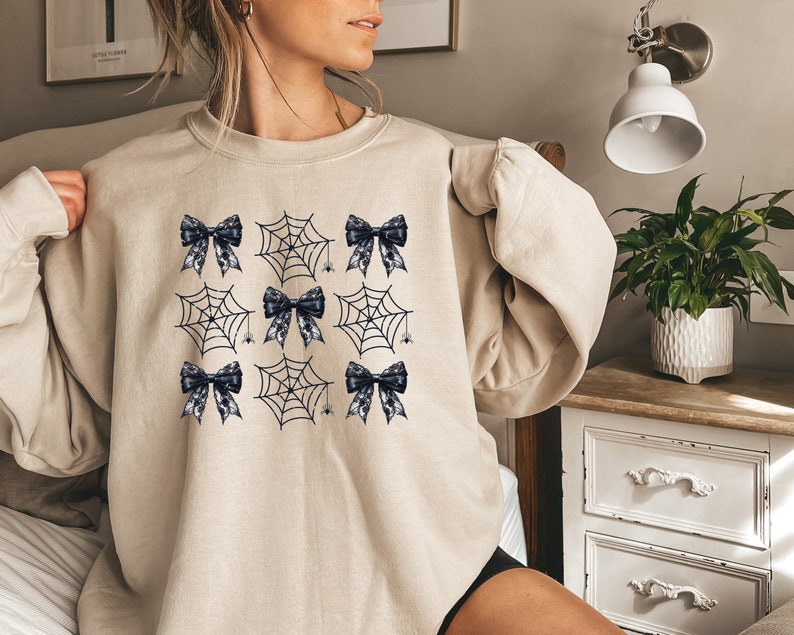Puede incluir: Una sudadera beige con un patr&oacute;n de telara&ntilde;a negra y blanca y lazos negros.
