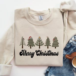 May include: A cream-colored sweatshirt with a festive design of five Christmas trees with red and green accents. The text "Merry Christmas" is printed in black below the trees.