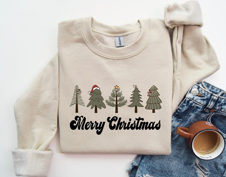 May include: A cream-colored sweatshirt with a festive design of five Christmas trees with red and green accents. The text "Merry Christmas" is printed in black below the trees.