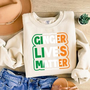 May include: A cream-colored sweatshirt with the text "Ginger Lives Matter" in green, white, and orange letters. The text is arranged in a rectangle with a green, white, and orange color scheme, resembling the Irish flag.