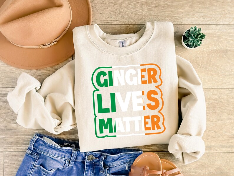 May include: A cream-colored sweatshirt with the text "Ginger Lives Matter" in green, white, and orange letters. The text is arranged in a rectangle with a green, white, and orange color scheme, resembling the Irish flag.