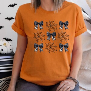 Puede incluir: Camiseta naranja con un dise&ntilde;o de telara&ntilde;a y lazo en blanco y negro. El dise&ntilde;o presenta m&uacute;ltiples telara&ntilde;as con un lazo en cada telara&ntilde;a.