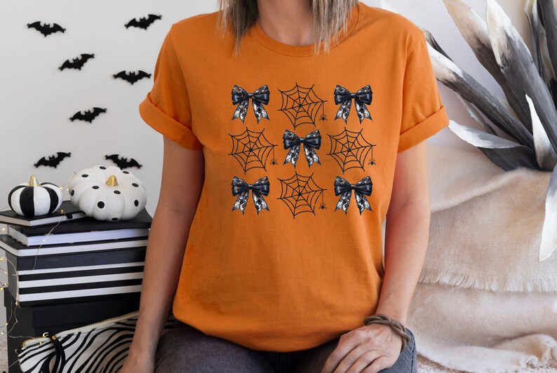 Puede incluir: Camiseta naranja con un dise&ntilde;o de telara&ntilde;a y lazo en blanco y negro. El dise&ntilde;o presenta m&uacute;ltiples telara&ntilde;as con un lazo en cada telara&ntilde;a.