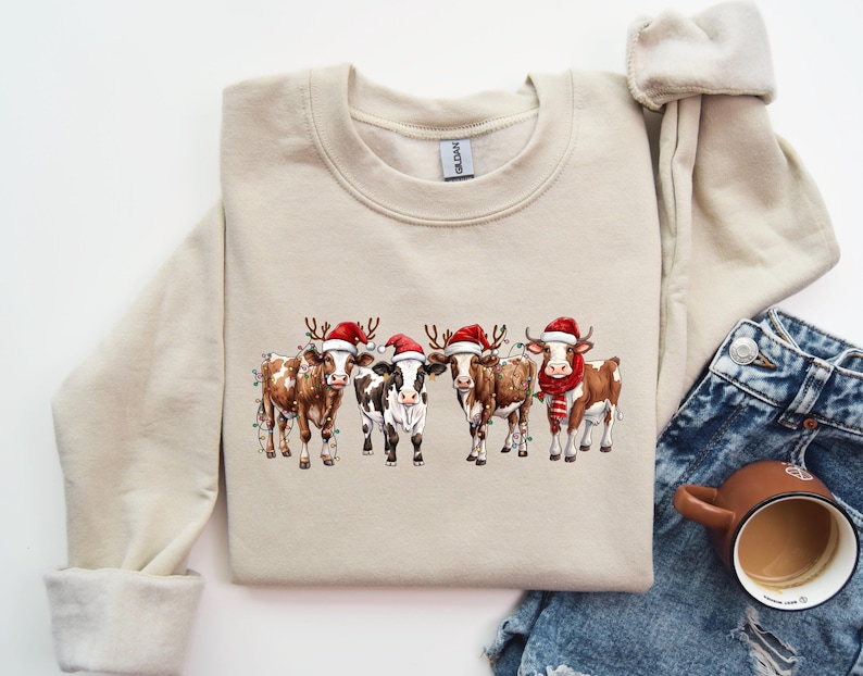 May include: A beige sweatshirt with a Christmas-themed design featuring four cows wearing Santa hats and reindeer antlers. The cows are adorned with Christmas lights and are standing in a row.