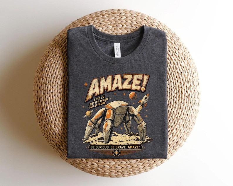 May include: A dark heather gray t-shirt featuring a graphic of a robotic crab on a lunar surface with a rocket and planets. The text reads AMAZE! LIFE IS BETTER WHEN YOU EXPLORE and BE CURIOUS. BE BRAVE. AMAZE! in a retro, distressed orange and cream font.