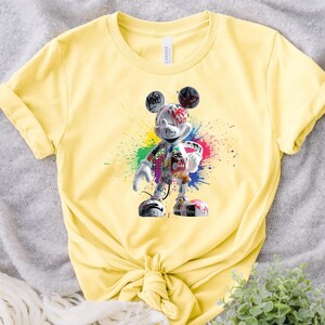 May include: Pale yellow t-shirt with a graphic of Mickey Mouse in a graffiti style. The design incorporates colourful paint splatters and text on the character. The t-shirt is tied at the waist.
