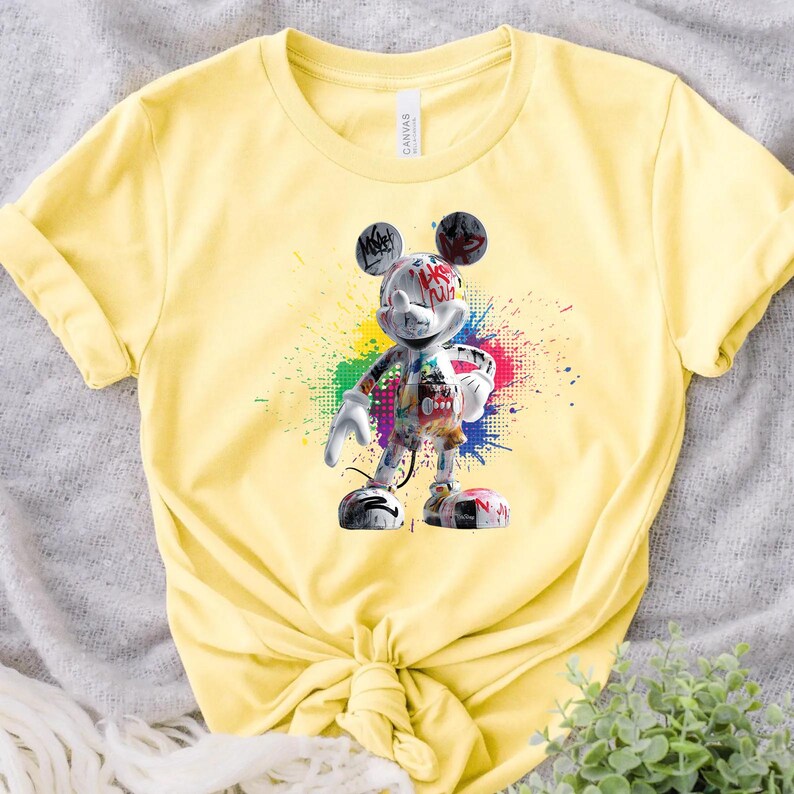May include: Pale yellow t-shirt with a graphic of Mickey Mouse in a graffiti style. The design incorporates colourful paint splatters and text on the character. The t-shirt is tied at the waist.