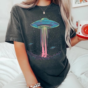 May include: Dark grey t-shirt with a vibrant graphic of a UFO beaming light downwards. The design includes a starry sky and other UFOs. The t-shirt has a relaxed fit.