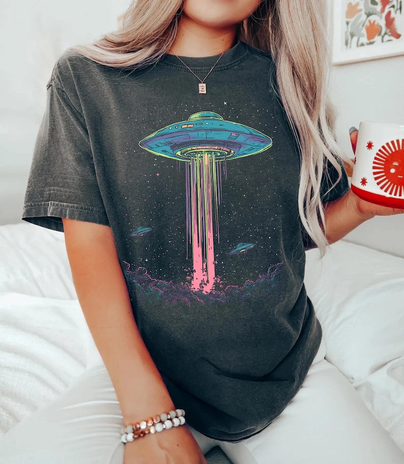 May include: Dark grey t-shirt with a vibrant graphic of a UFO beaming light downwards. The design includes a starry sky and other UFOs. The t-shirt has a relaxed fit.