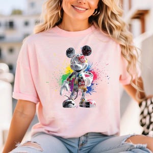 May include: A pale pink t-shirt with a graffiti-style Mickey Mouse design. The Mickey Mouse figure is adorned with colourful paint splatters and graffiti art. The t-shirt has short sleeves and a loose fit.